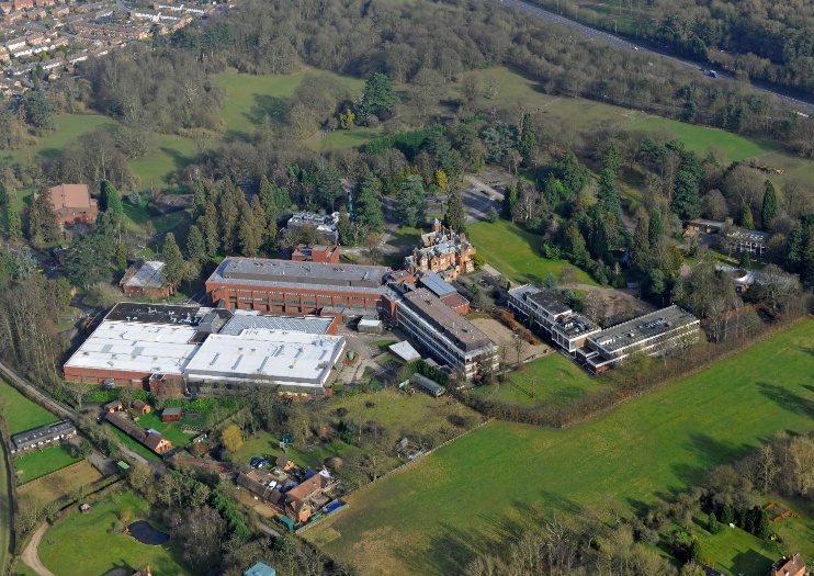 Aerial view of industrial complex with white-roofed warehouses, brick buildings, and multi-storey structures surrounded by green fields and trees.