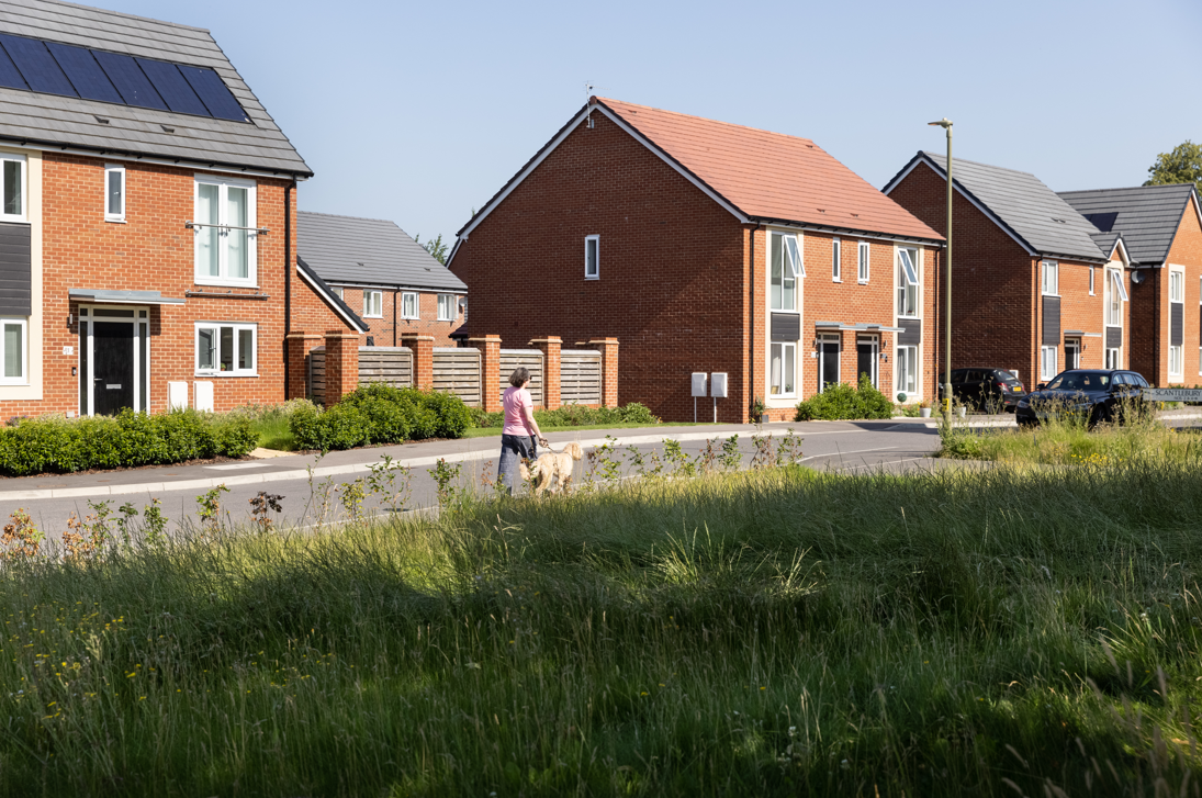 Modern brick housing estate with red-brown houses, solar panels, wooden fencing, and green grass verge in foreground.
