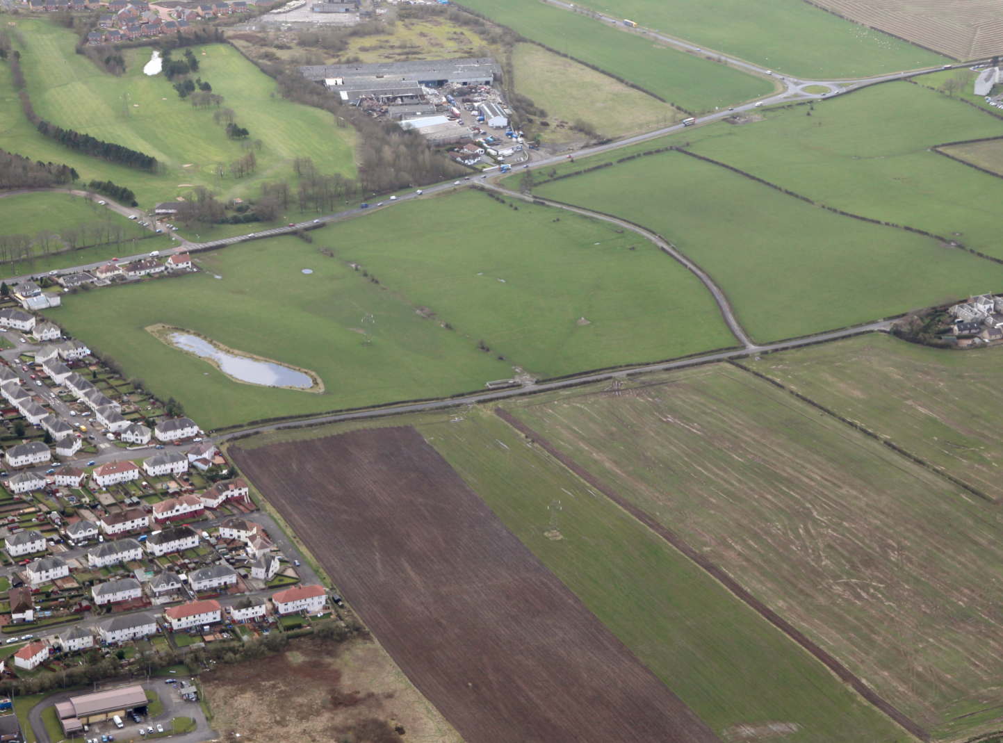 Aerial view of rural countryside with green fields, residential housing estate, curved roads, and industrial buildings in distance.