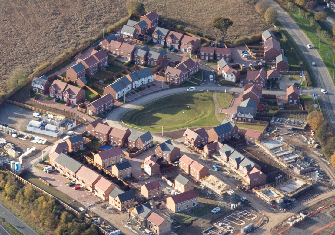 Aerial view of residential housing estate with red-tiled roofs arranged around central green space, surrounded by roads and fields.