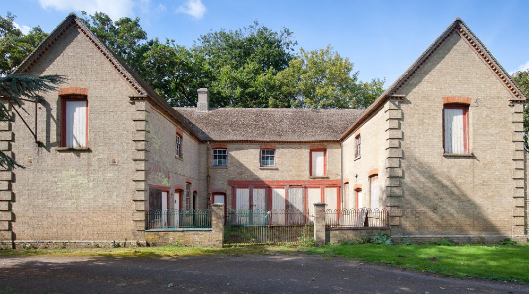 Modern residential complex with cream-coloured stone walls, red-framed windows, and terracotta roof tiles under blue sky with trees.