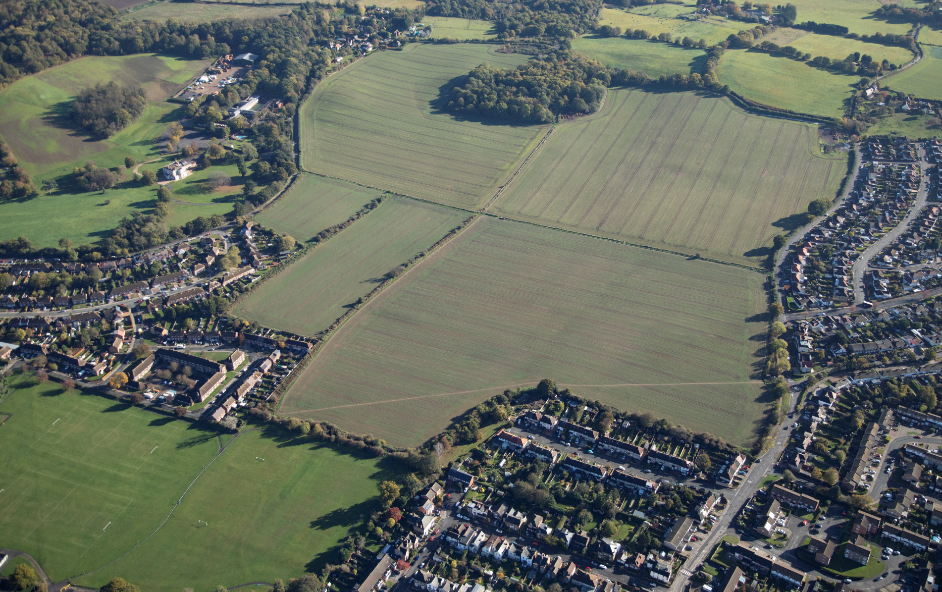 Aerial view of suburban housing estates surrounding large green agricultural fields with hedgerow boundaries and scattered trees.