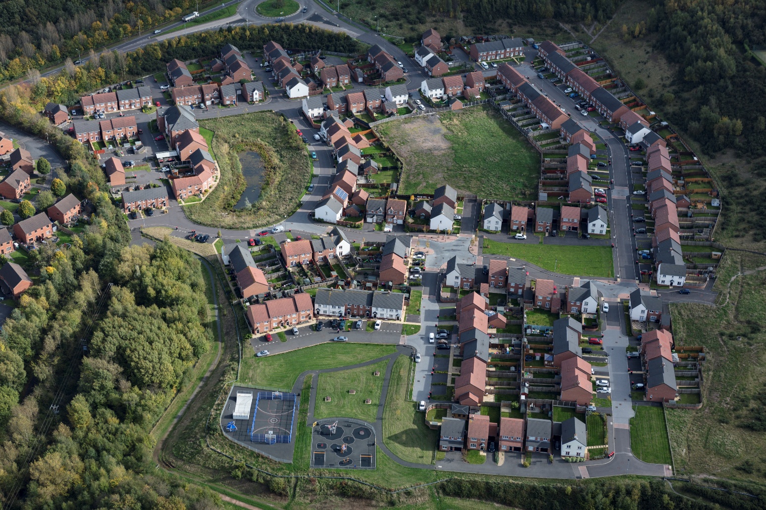 Aerial view of residential housing estate with red-roofed houses, green spaces, car parks, and surrounding woodland.