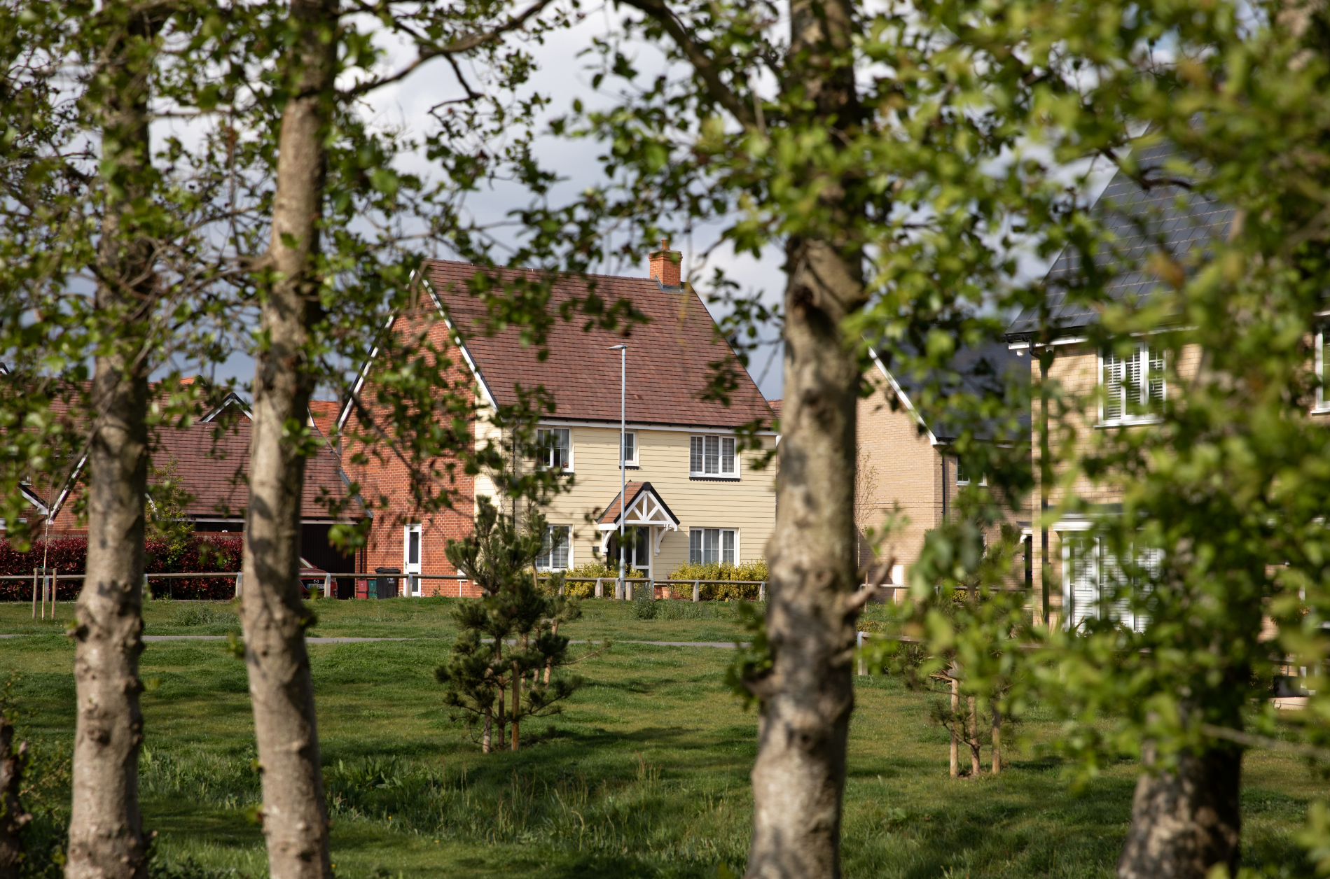 Red-brick house with tiled roof viewed through green tree branches and foliage, with lawn and neighbouring buildings visible.