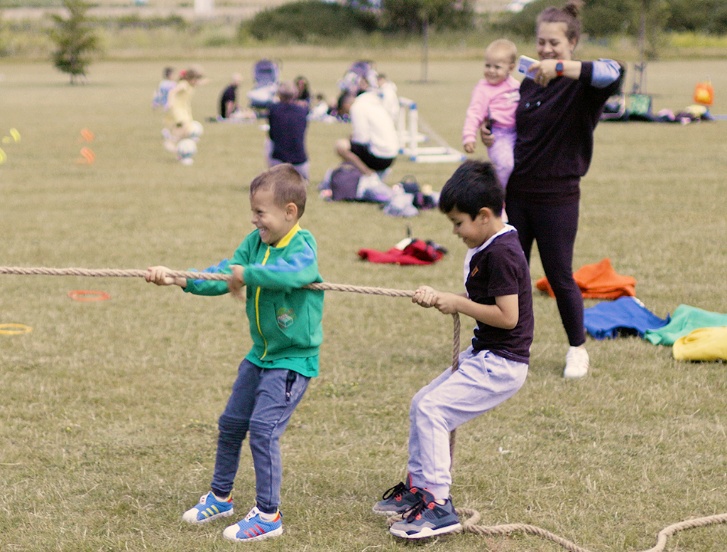 Children playing tug-of-war on grass field with families watching in background during outdoor event