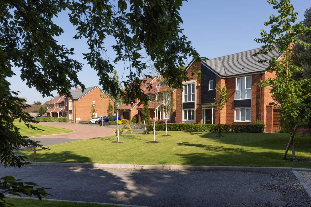 Modern residential street with red brick houses, manicured green lawns, and mature trees under blue sky with white clouds.