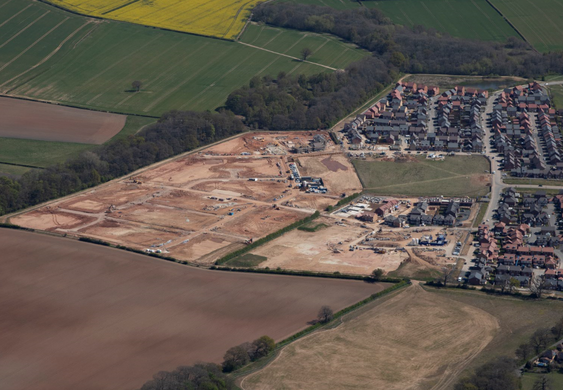 Aerial view of housing development construction site with brown earthworks adjacent to existing residential streets and green fields.
