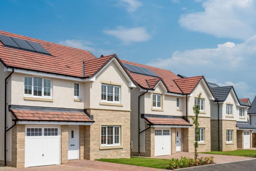 Row of modern two-storey houses with cream render walls, red clay tile roofs, white garage doors, and small front gardens under blue sky.
