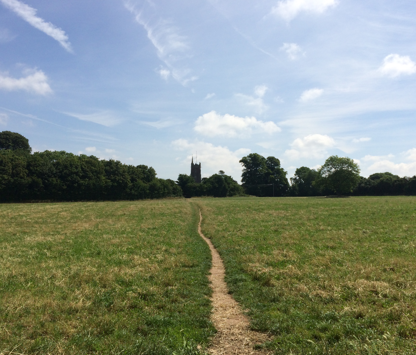 Dirt path crosses green field towards church tower, with trees lining horizon under blue sky with white clouds.