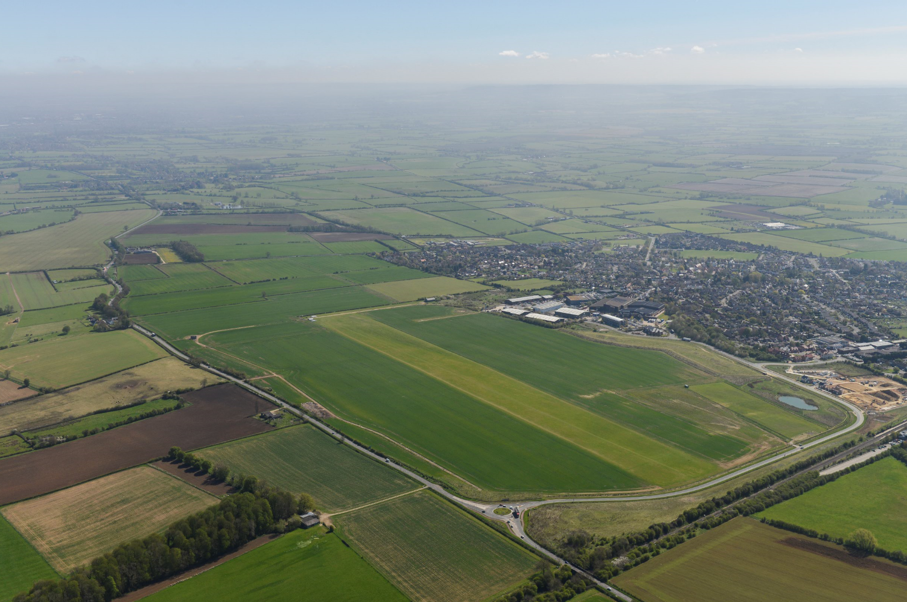 Aerial view of airport runway surrounded by green fields and farmland, with town visible in background under cloudy sky.