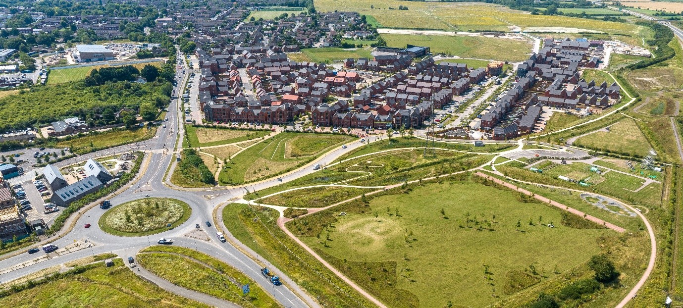 Aerial view of a new housing development beside a large green park, with winding footpaths, roads, a roundabout, fields and surrounding suburban neighbourhood.